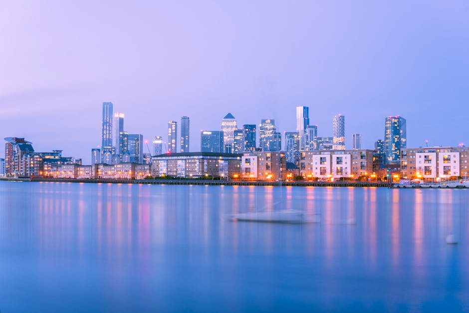 A panoramic view of Canary Wharf's city skyline at dusk, featuring modern skyscrapers with illuminated windows and sleek glass facades reflecting on the still waters of the Thames River. The scene inc