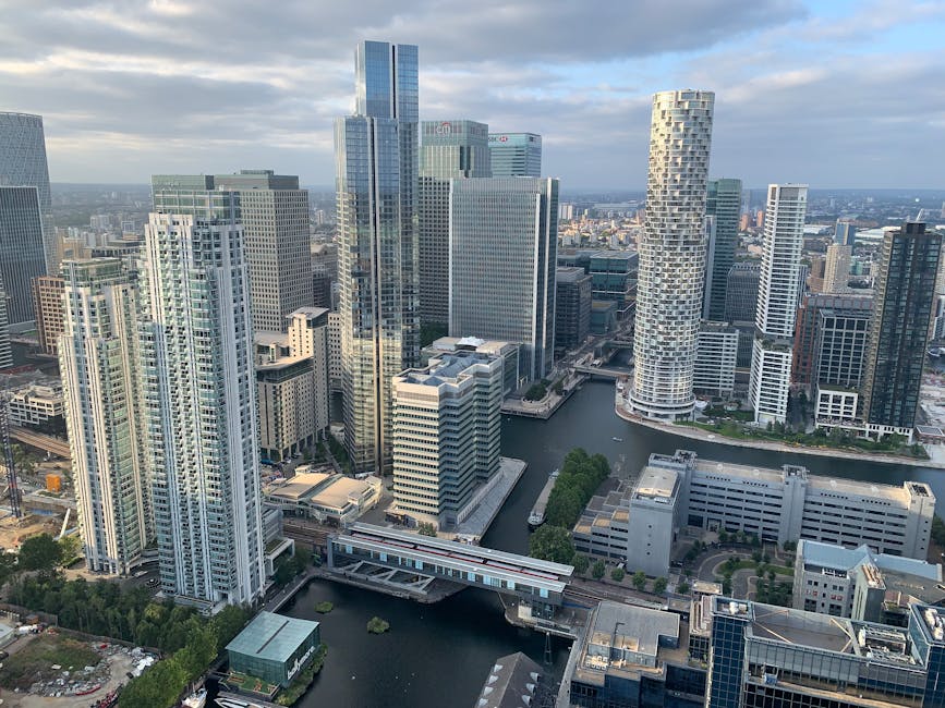 A panoramic view of Canary Wharf's city skyline at dusk, featuring modern skyscrapers with illuminated windows and sleek glass facades reflecting on the still waters of the Thames River. The scene inc