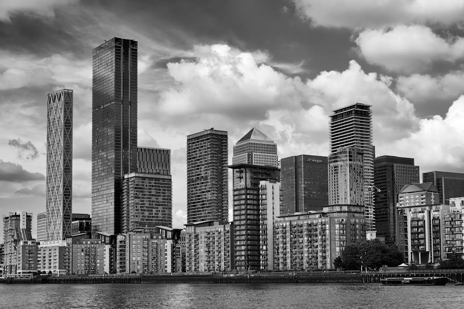 Black and white aerial photograph of a modern urban area in Canary Wharf, showcasing a cluster of high-rise office buildings with glass facades reflecting light. The foreground features a circular lan
