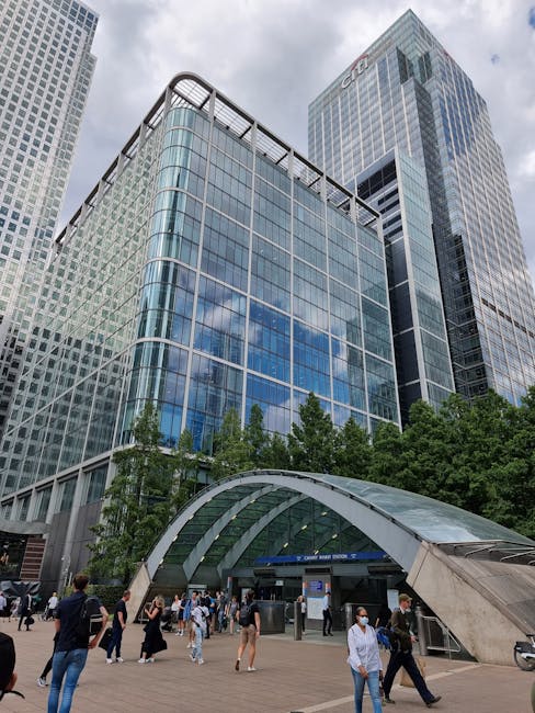 The image showcases a spacious interior with a large floor-to-ceiling glass facade, providing a clear view of a modern cityscape with tall office buildings under a bright blue sky. The glass panels ar