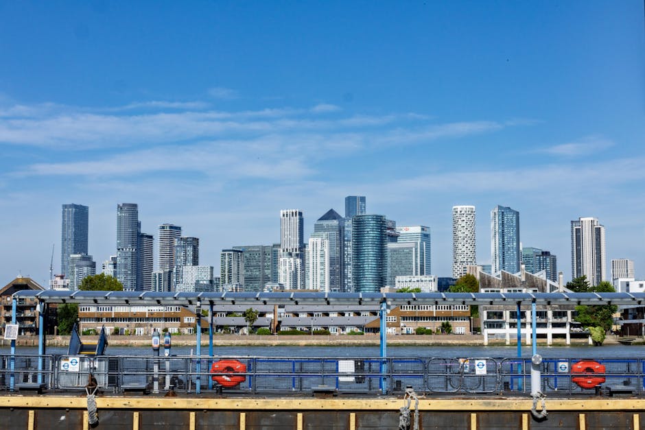 A cluster of modern, multi-level brick residential buildings featuring sloped roofs, numerous balconies with white railings, and large windows, situated along a waterway. In the background, a city sky